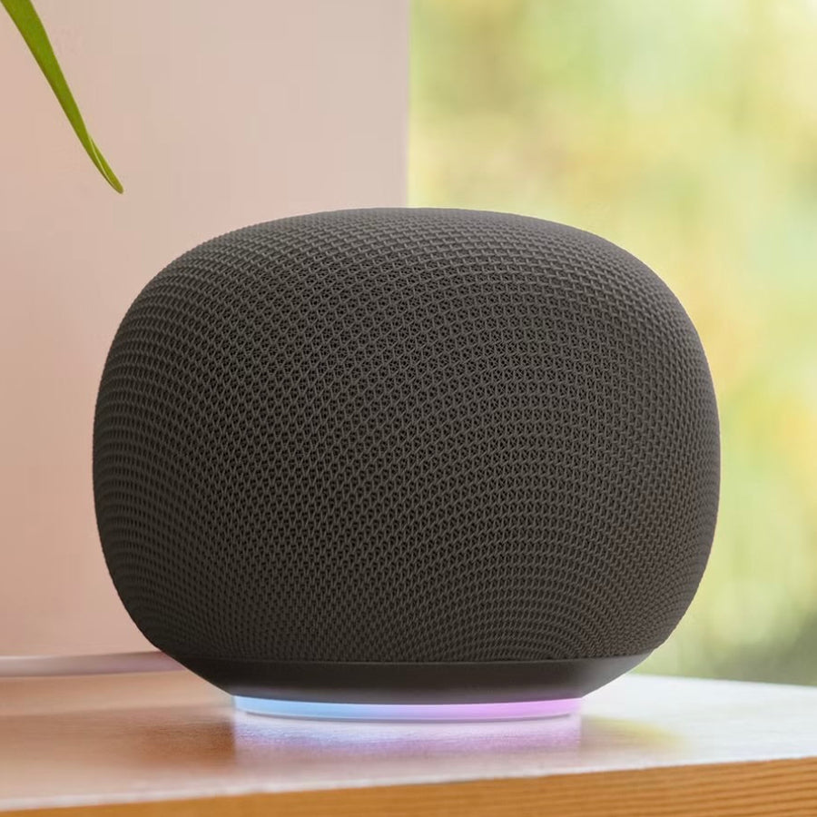 A dark gray spherical smart speaker with a textured fabric mesh cover sits on a wooden surface, casting a soft purple and blue light glow. A red ceramic vase with green leaves is visible on the left, and a softly blurred window background with green foliage is on the right.