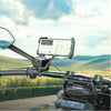 A black smartphone mount attached to a motorcycle handlebar, holding a phone with a navigation screen. The motorcycle dashboard and side mirror are visible, with a blurred outdoor landscape in the background.
