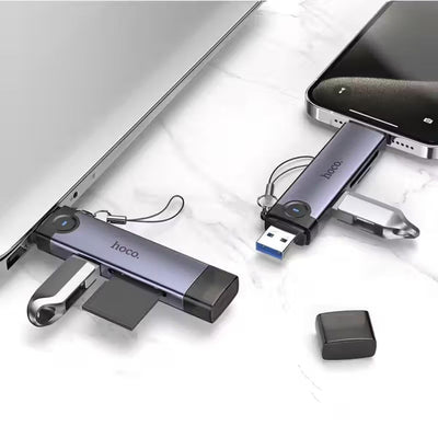 Two Hoco. USB flash drives in a metallic purple-gray finish are shown plugged into a laptop and smartphone on a white marble surface, with one drive's cap removed and placed beside it.