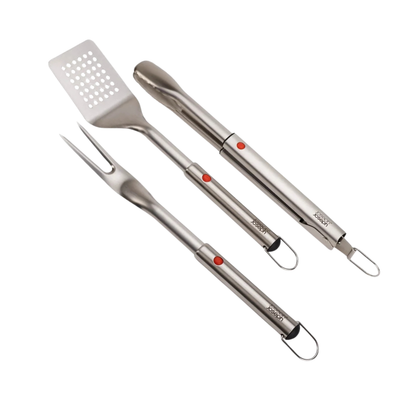 Joseph Joseph GrillOut 3-piece BBQ set featuring stainless-steel turner, tongs, and fork on a wooden surface.