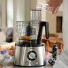 A Philips food processor with a stainless steel base and clear bowl is shown on a kitchen counter, with a person pouring water into the feeder hopper. Eggs, flour, and kitchen items are visible in a softly blurred kitchen setting.