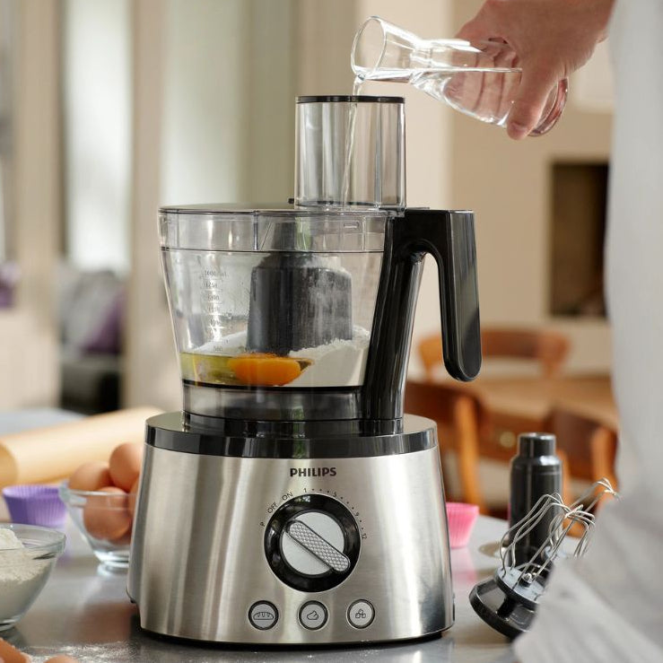 A Philips food processor with a stainless steel base and clear bowl is shown on a kitchen counter, with a person pouring water into the feeder hopper. Eggs, flour, and kitchen items are visible in a softly blurred kitchen setting.