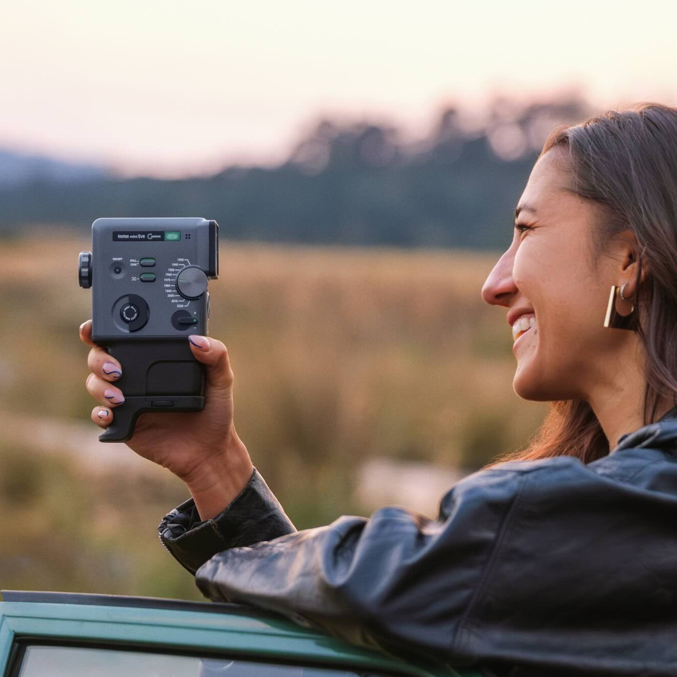A woman in a black leather jacket holds a black handheld device with dials and buttons, smiling as she looks out over a blurred outdoor landscape at sunset.