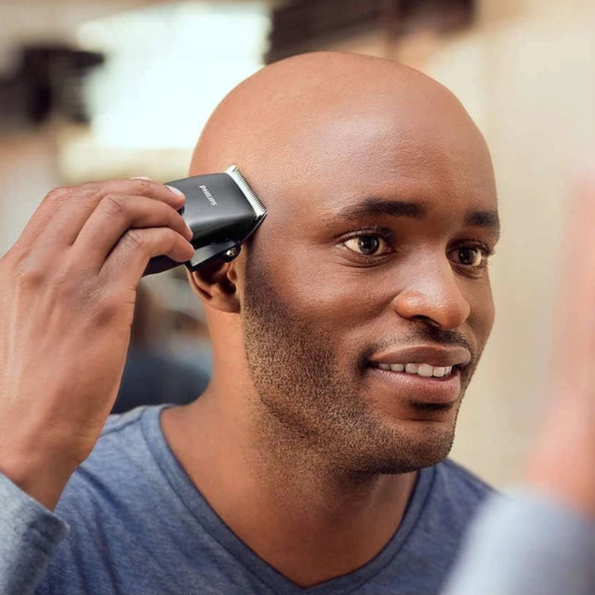 A man with a shaved head uses a black and silver electric clipper in a grooming setting, with a blurred reflection visible in the mirror.