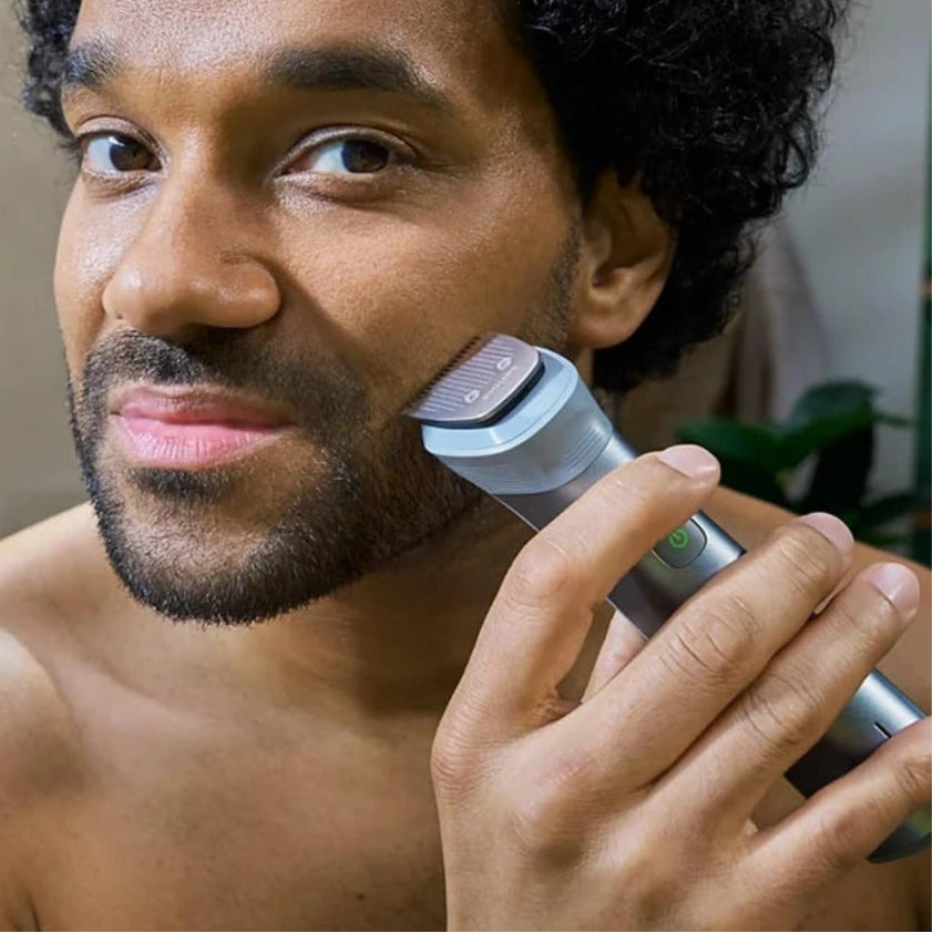 A man with curly black hair and a beard shaves his face using a silver and gray electric trimmer in a bathroom setting.