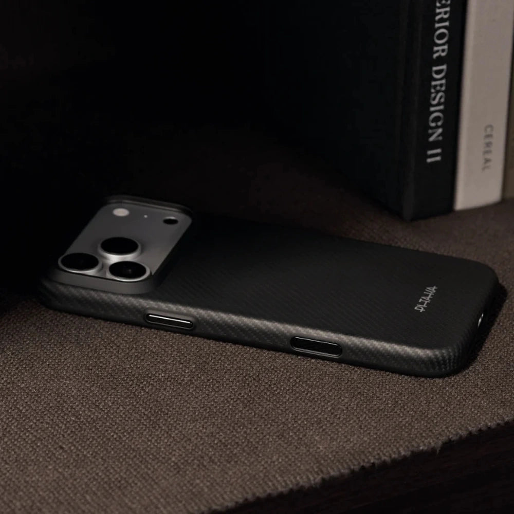 A black carbon-fiber-textured phone case on a smartphone, resting on a dark textured surface with books in the background.